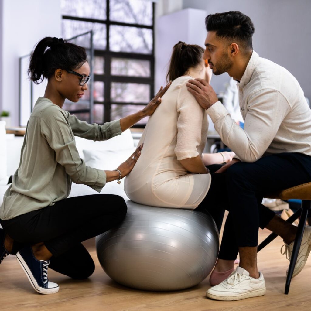 A man and woman sit on an exercise ball, engaged in a doula training session focused on supporting families.