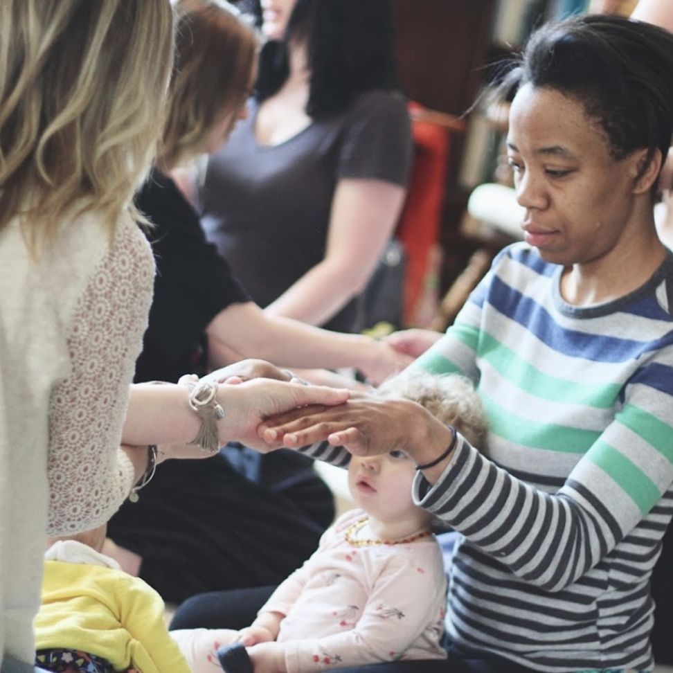 A woman holds a baby while another woman gently holds her hand, conveying a moment of support and connection.